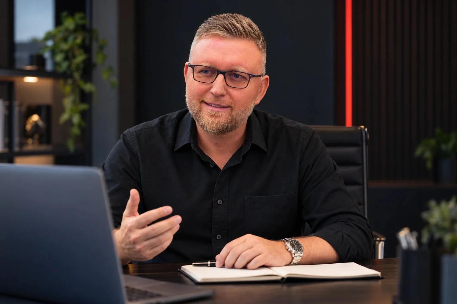 Sascha Frenz im schwarzen Hemd am Schreibtisch mit Laptop und Notizbuch.